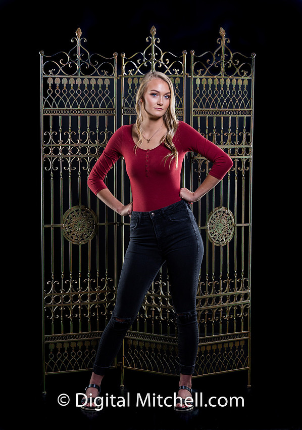 photograph of a high school senior posing in a red top and blue genes on a black background in front of a metal room divider