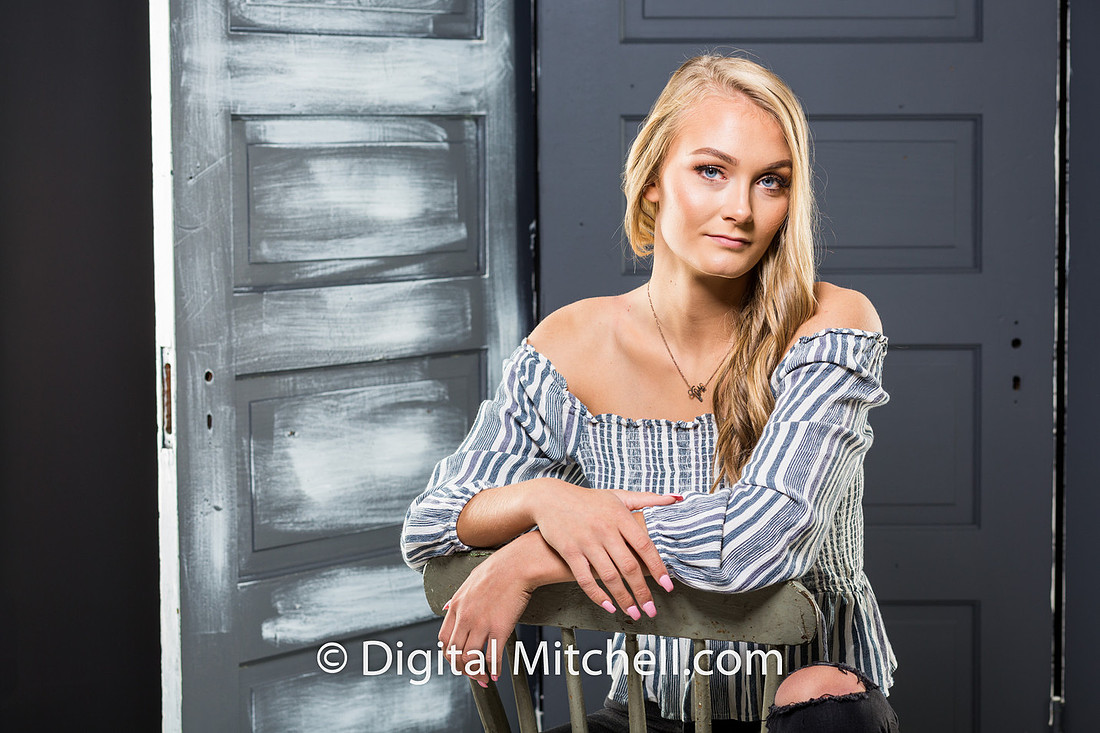 photograph of a high school senior posing in a gray top and black genes in front of a gray set of doors 